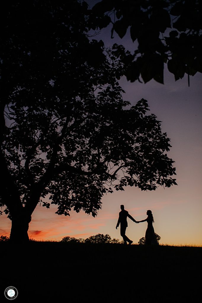 groom leads bride across a hill with a golden sunset the couple and trees are silhouetted against the sunset sky