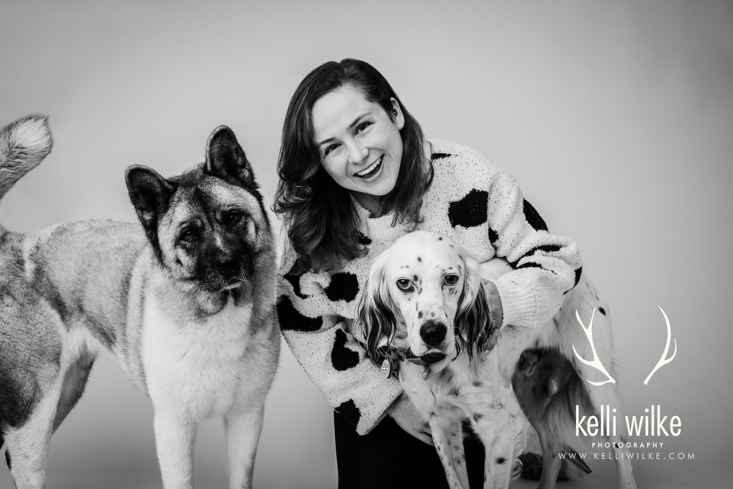A black and white posed portrait of a woman in a white sweater with two black and white dogs during a pet portrait session with Kelli Wilke Photography in Wilmington, Delaware. 