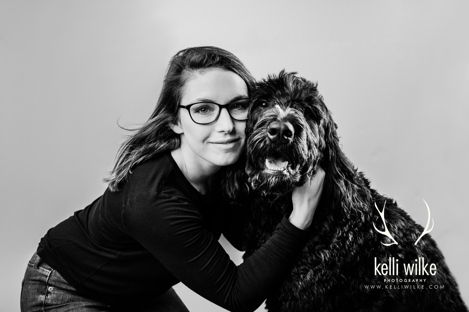 A black and white picture of a woman in glasses snuggling a large, shaggy, black dog during a pet portrait session in Wilmington, Delaware by Kelli Wilke Photography. 