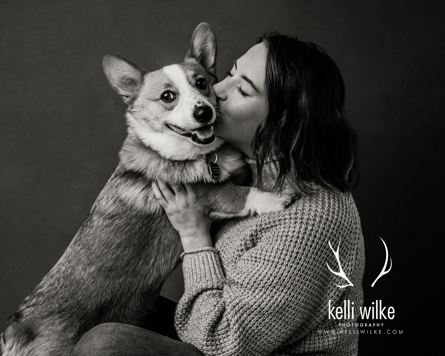 A black and white picture of a woman kissing a corgi on the cheek, the corgi smiling at the camera during a pet portrait session by Kelli Wilke Photography in Wilmington, Delaware. 