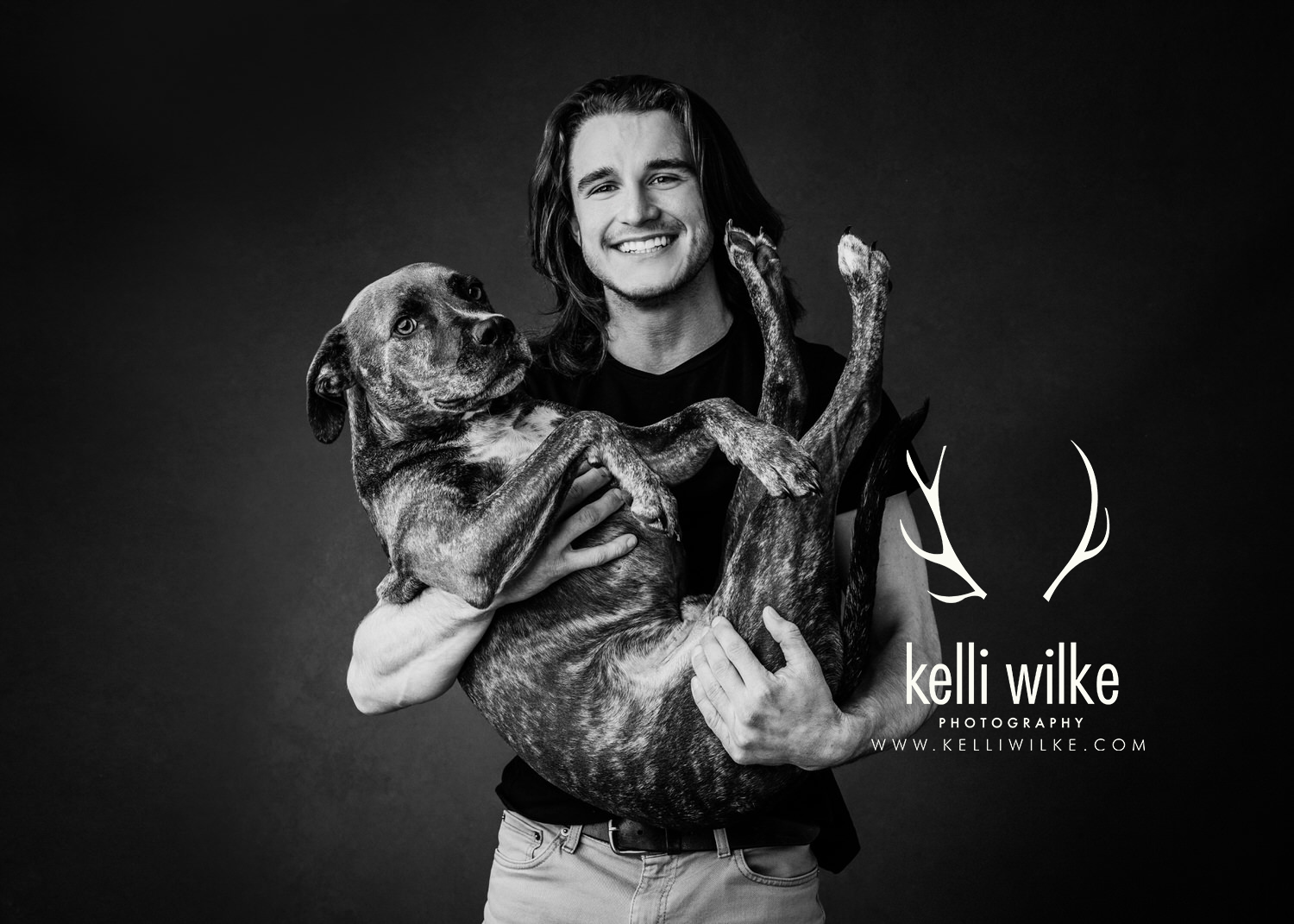 A black and white picture of a man with long hair holding a boxer in his arms, both smiling at the camera during a pet portrait session by Kelli Wilke Photography in Wilmington, Delaware. 