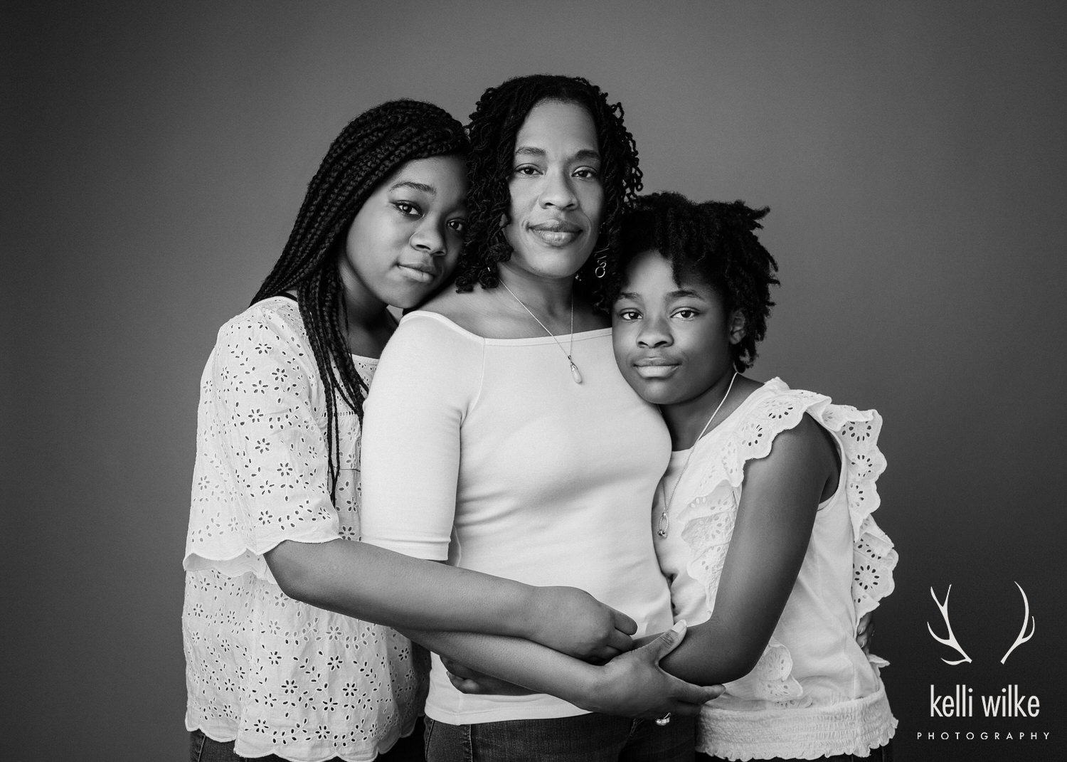 A black and white portrait of a mother with her two daughters, their arms wrapped around each other taken by Kelli Wilke Photography in Wilmington, Delaware. 
