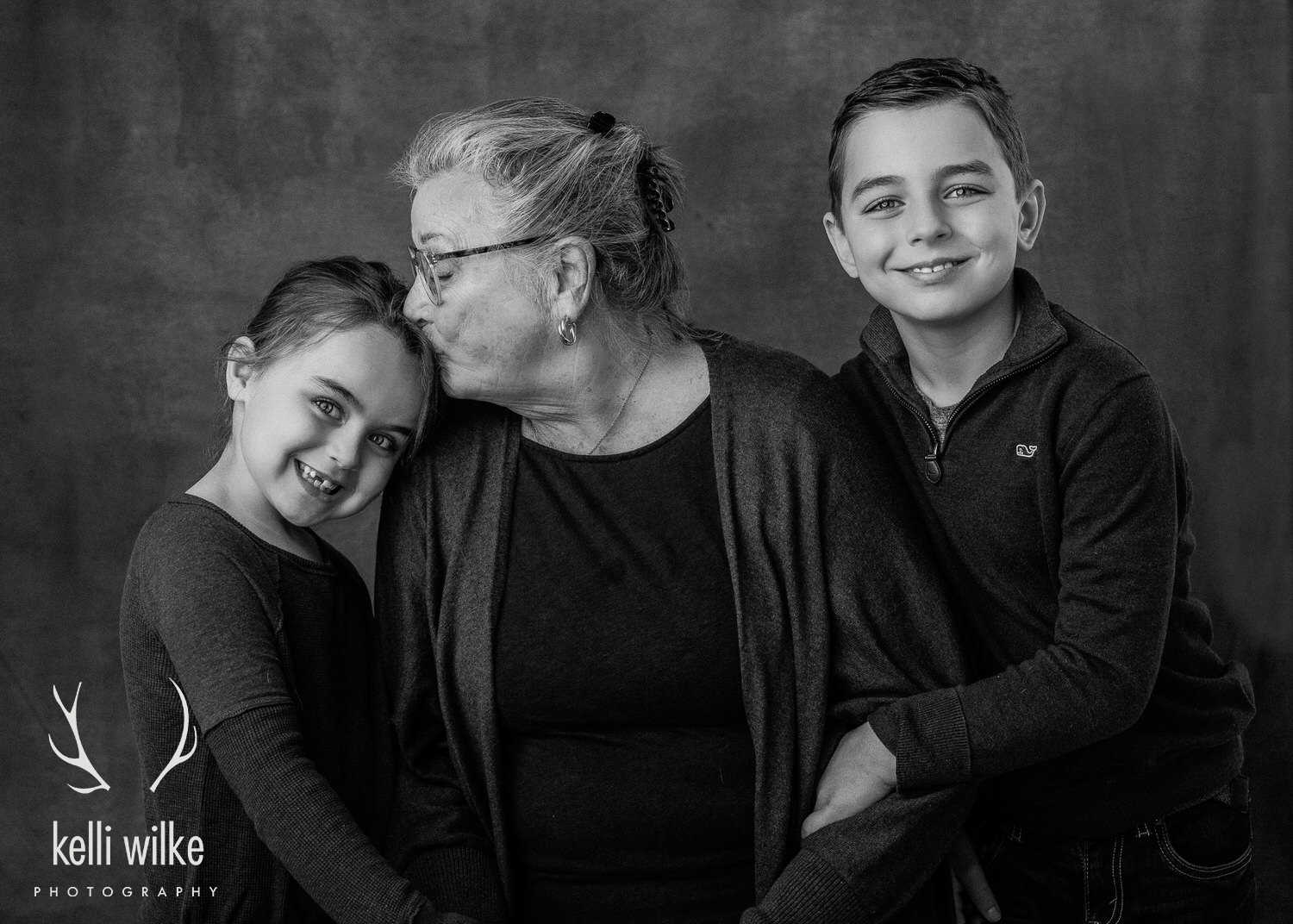 A black and white studio portrait of a grandmother and her two grandchildren; the grandmother kissing her granddaughter on her forehead taken by Kelli Wilke Photography in Wilmington, DE. 