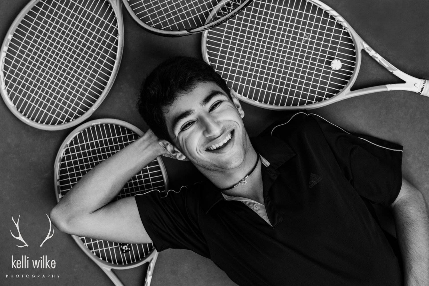 A black and white picture of a teenage boy laying on the ground on top of tennis rackets, smiling up at the camera, photographed by Kelli Wilke Photography. 