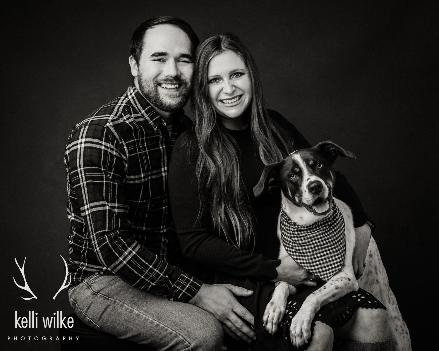 A black and white picture of a couple with their dog, all smiling at the camera. Photographed by Kelli Wilke Photography in Wilmington, Delaware. 