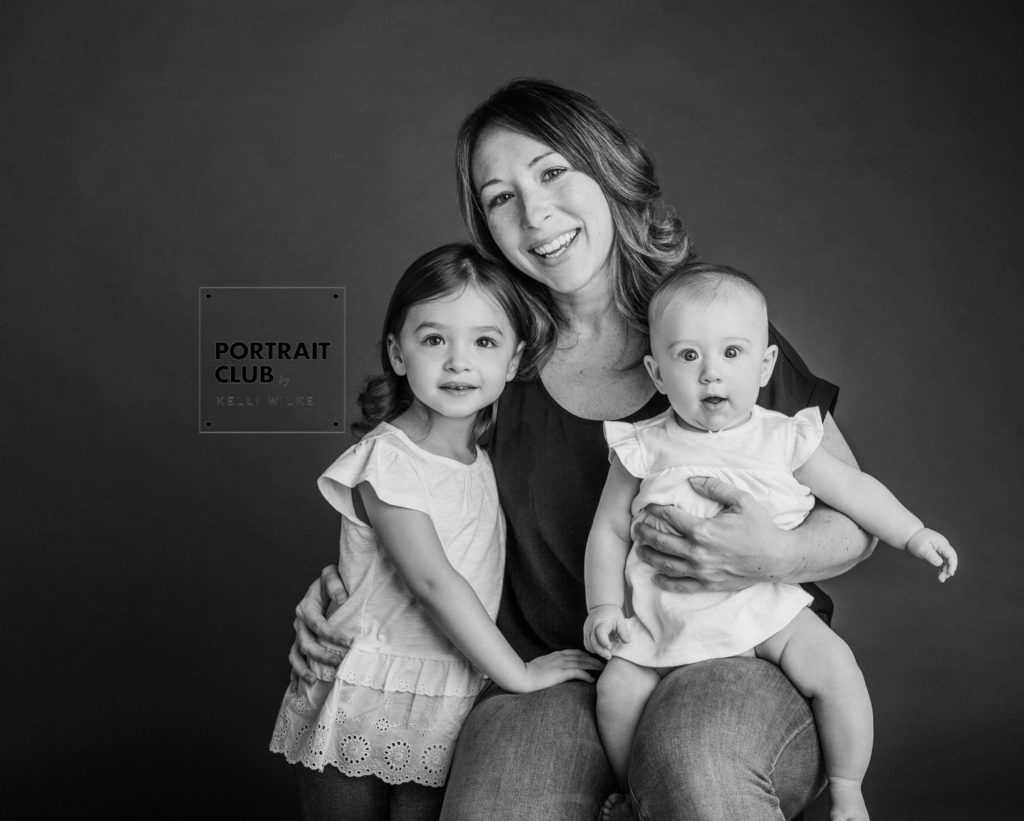 A candid black and white picture of a woman in a dark tee-shirt holding two little girls in white dresses during a studio portrait session in Wilmington Delaware by Portrait Club by Kelli Wilke. 