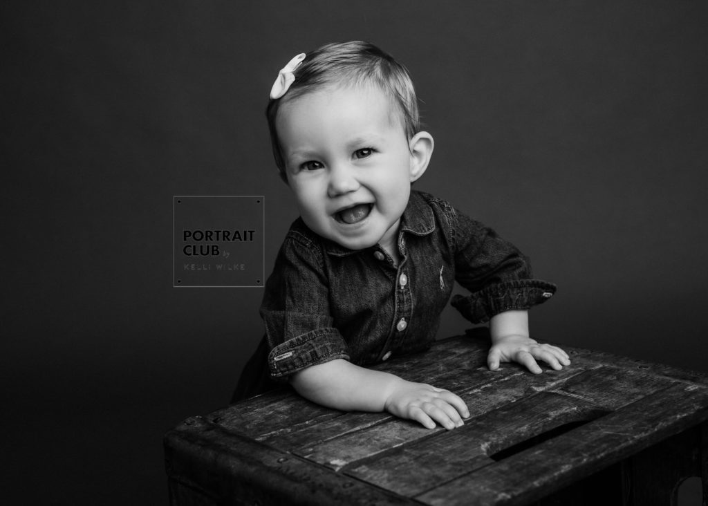 A candid black and white picture of a baby girl with a bow in her hair smiling and looking at the camera during a studio portrait session with Portrait Club by Kelli Wilke in Wilmington, Delaware. 