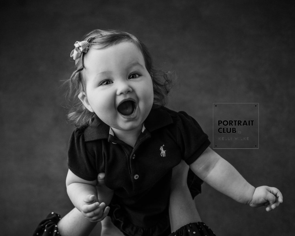 A candid black and white picture of a baby girl with a bow in her hair smiling and looking at the camera during a studio portrait session with Portrait Club by Kelli Wilke in Wilmington, Delaware. 