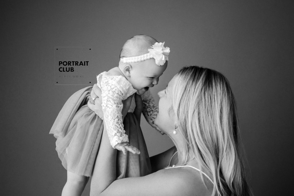 A candid picture of a woman with long blonde hair holding up a baby girl in a ruffly dress and hair bow, both smiling and laughing at each other during a studio portrait session with Portrait Club by Kelli Wilke Photography in Wilmington, Delaware. 