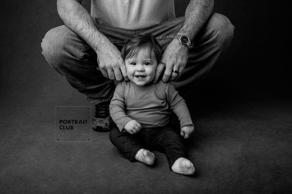 A candid picture of a baby smiling at the camera as his dad tickles his neck during a portrait session with Portrait Club by Kelli Wilke Photography in Wilmington, DE. 