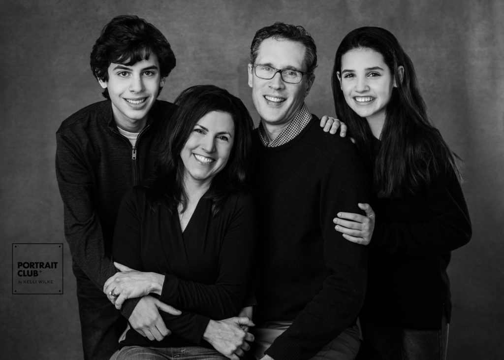 A formal portrait of a family in dark clothes smiling at the camera during a studio portrait session with Portrait Club by Kelli Wilke in Wilmington, DE.  
