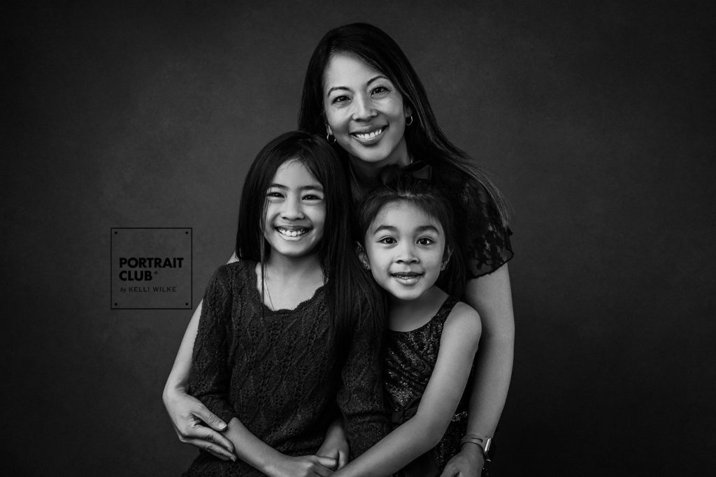 A formal portrait of a mom and her two little girls smiling at the camera during a family portrait session with Portrait Club by Kelli Wilke. 