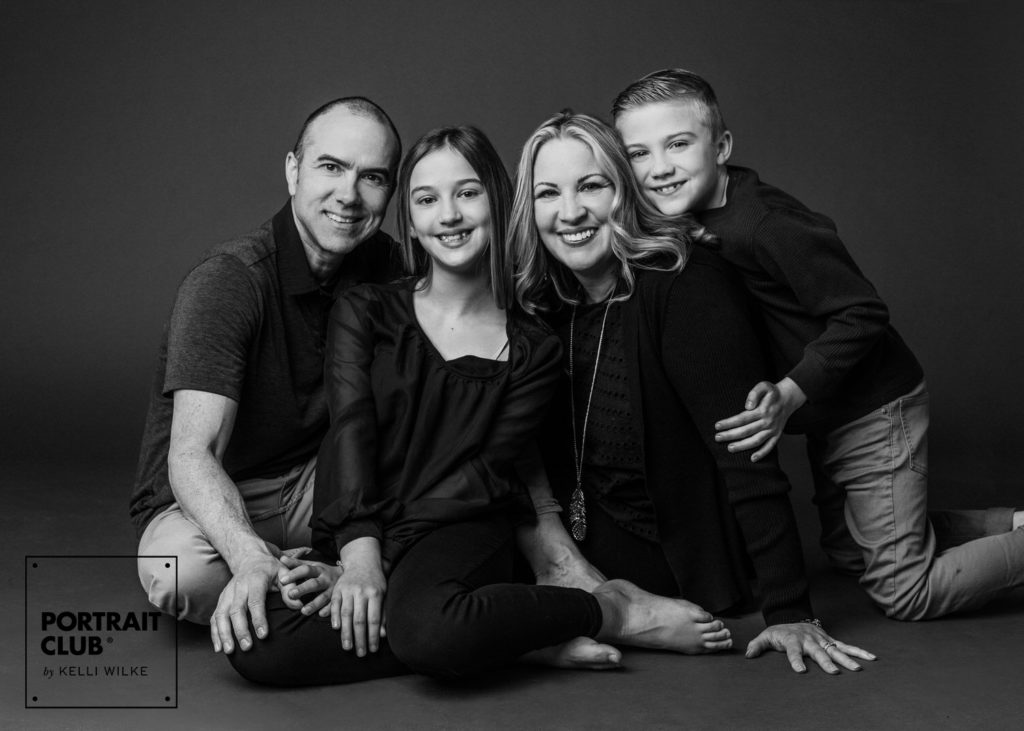 A candid black and white picture of a family sitting on the ground, smiling at the camera during a family photo session with Portrait Club by Kelli Wilke. 