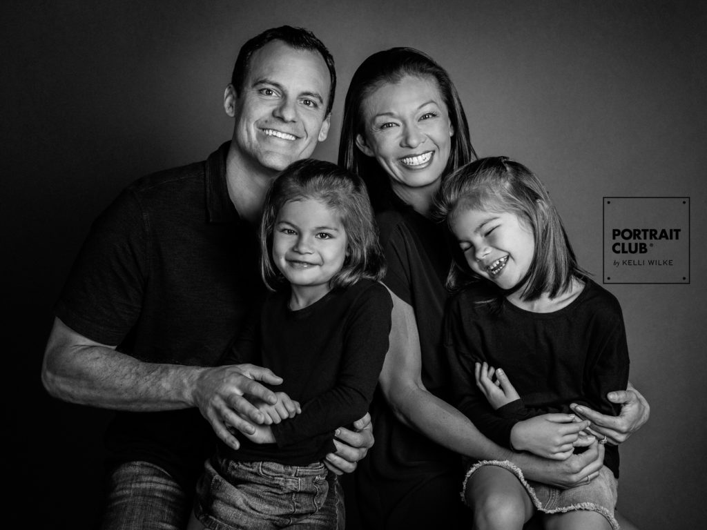 A candid picture of a family with two little girls smiling at the camera during a family photo session with Portrait Club by Kelli Wilke. 
